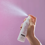 A hand holding POM Soothing Spray, spraying a fine mist against a soft purple background.